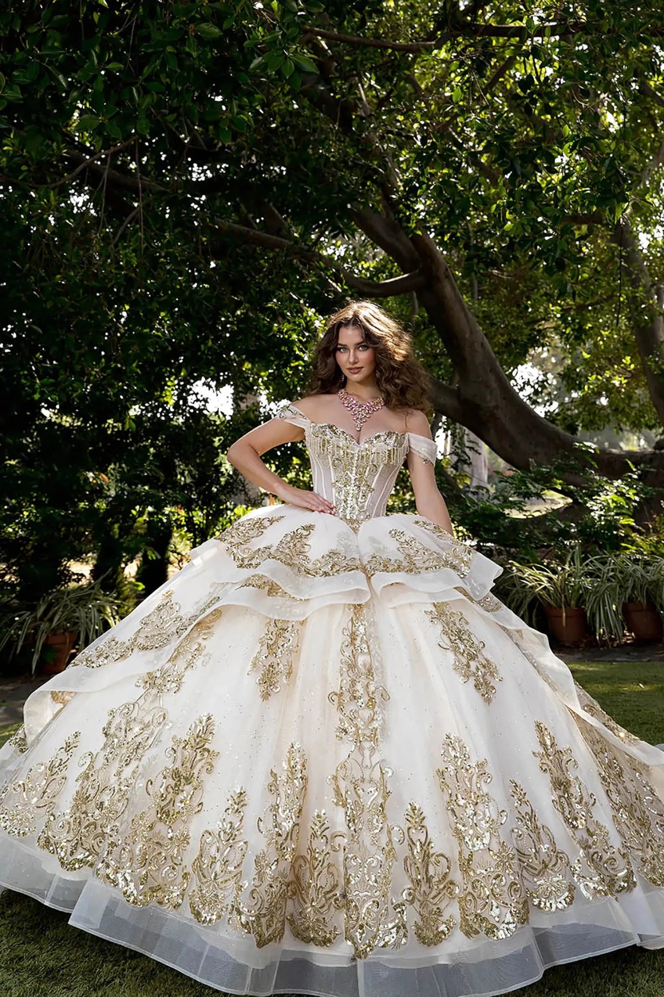 Woman wearing off-shoulder ball gown with sweetheart corset bodice, gold embroidery, layered tulle skirt, and lace-up back in an outdoor garden setting.