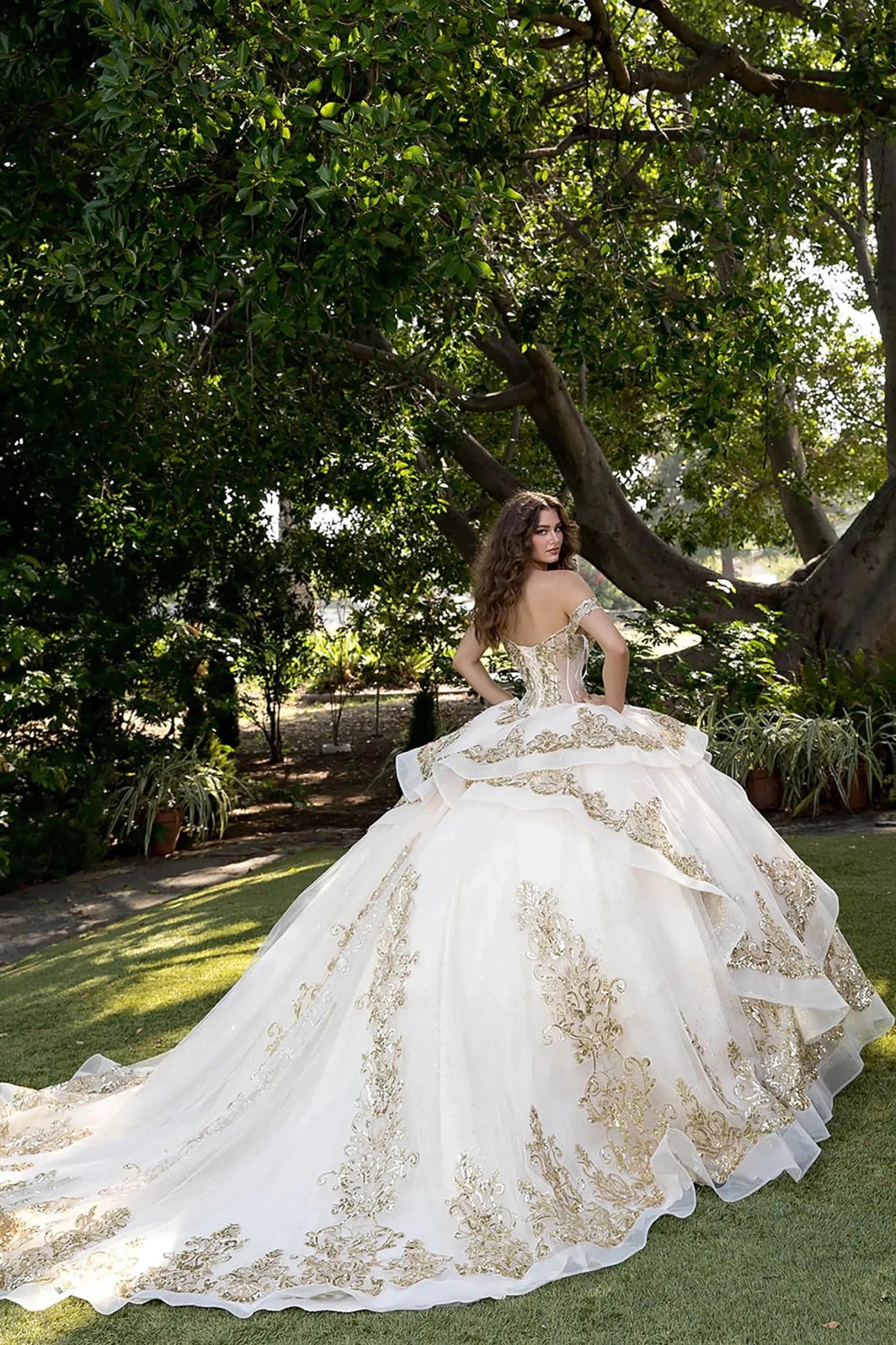 Model in champagne glitter tulle ball gown with lace-up corset and gold embroidery in outdoor garden.