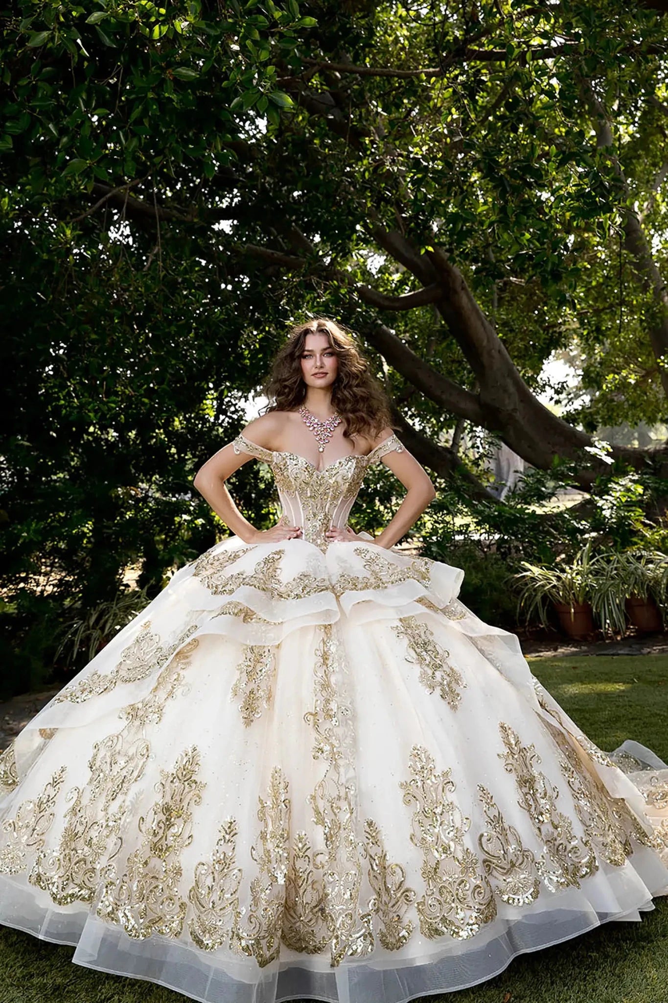 Front view of woman in champagne tulle ball gown with off-shoulder sleeves and gold appliqué corset outdoors.