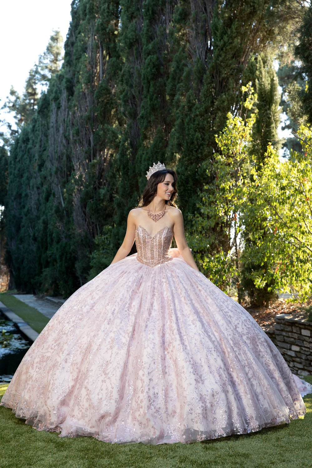 Woman in a pink lace dress with a tiara standing outdoors among trees.