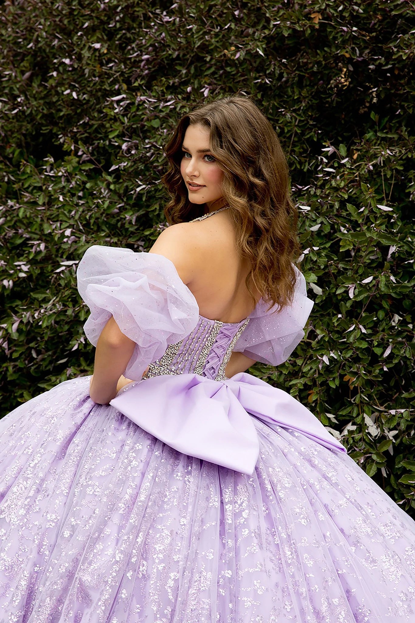 Woman in a lavender gown with puffed sleeves standing against a green hedge.