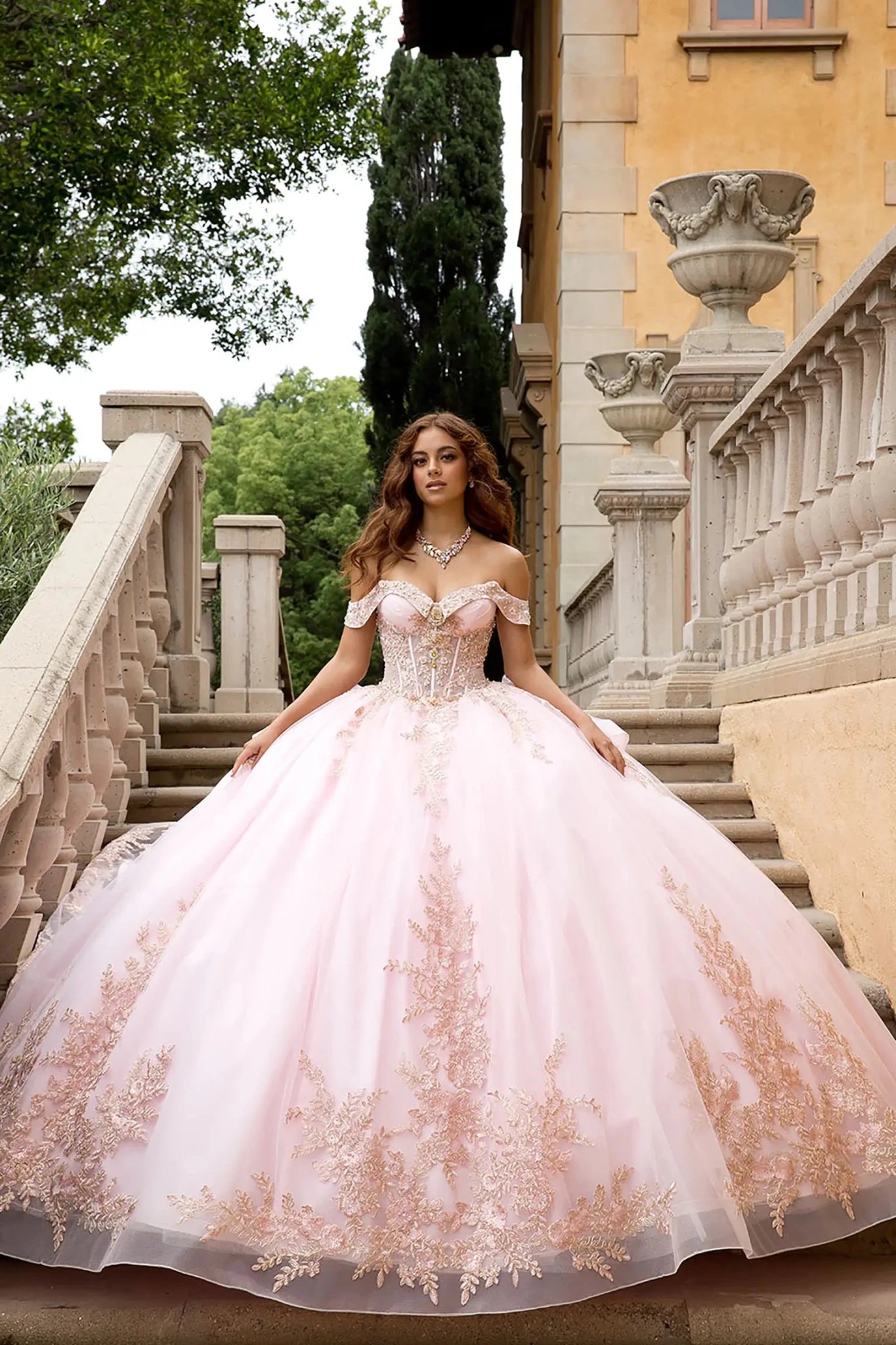 Lifestyle and Architectural Setting
A model wearing the pink PQ1082 Bella Princess Couture gown posing on a classical stone staircase. The image emphasizes the scale of the voluminous ball gown skirt, the sparkle of the rose gold lace, and the elegant off-the-shoulder silhouette against a Mediterranean-style architectural backdrop.