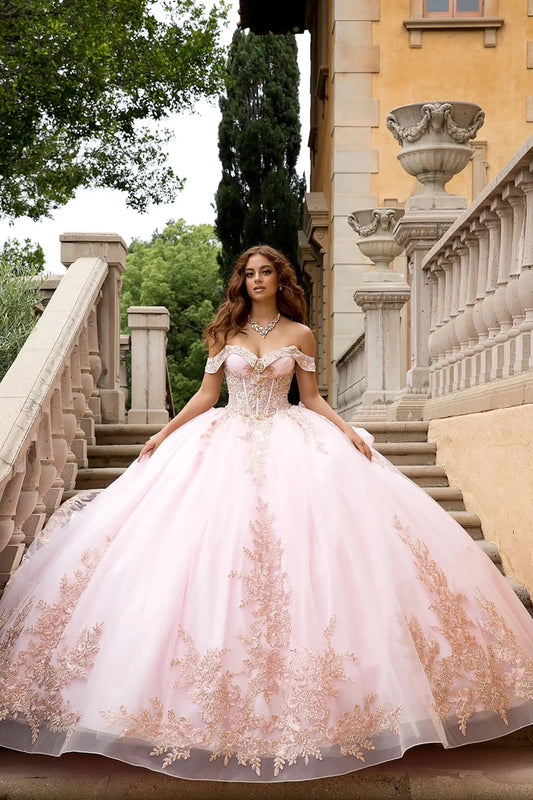 Lifestyle and Architectural Setting
A model wearing the pink PQ1082 Bella Princess Couture gown posing on a classical stone staircase. The image emphasizes the scale of the voluminous ball gown skirt, the sparkle of the rose gold lace, and the elegant off-the-shoulder silhouette against a Mediterranean-style architectural backdrop.