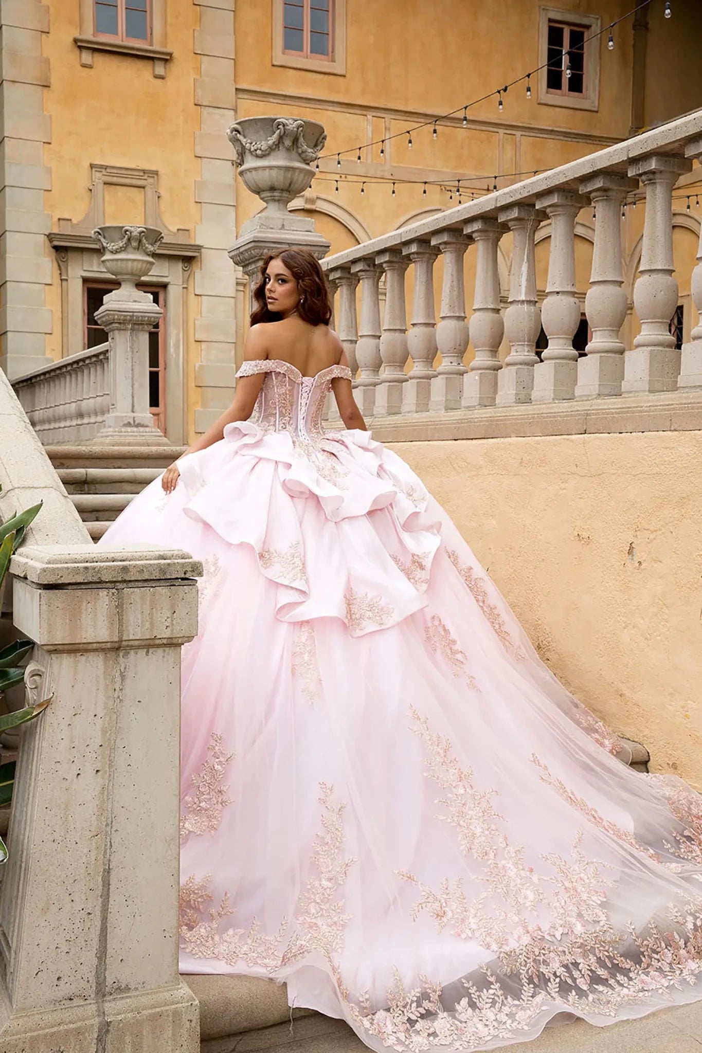 Rear editorial view showcasing the sculptural tiered satin ruffles and the adjustable lace-up corset back of the gown. The image captures the full length of the cathedral-style train adorned with shimmering rose gold floral lace motifs on a stone balcony.