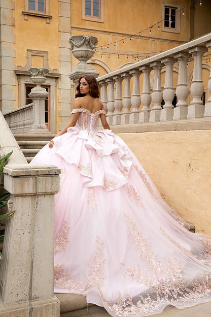 Rear editorial view showcasing the sculptural tiered satin ruffles and the adjustable lace-up corset back of the gown. The image captures the full length of the cathedral-style train adorned with shimmering rose gold floral lace motifs on a stone balcony.