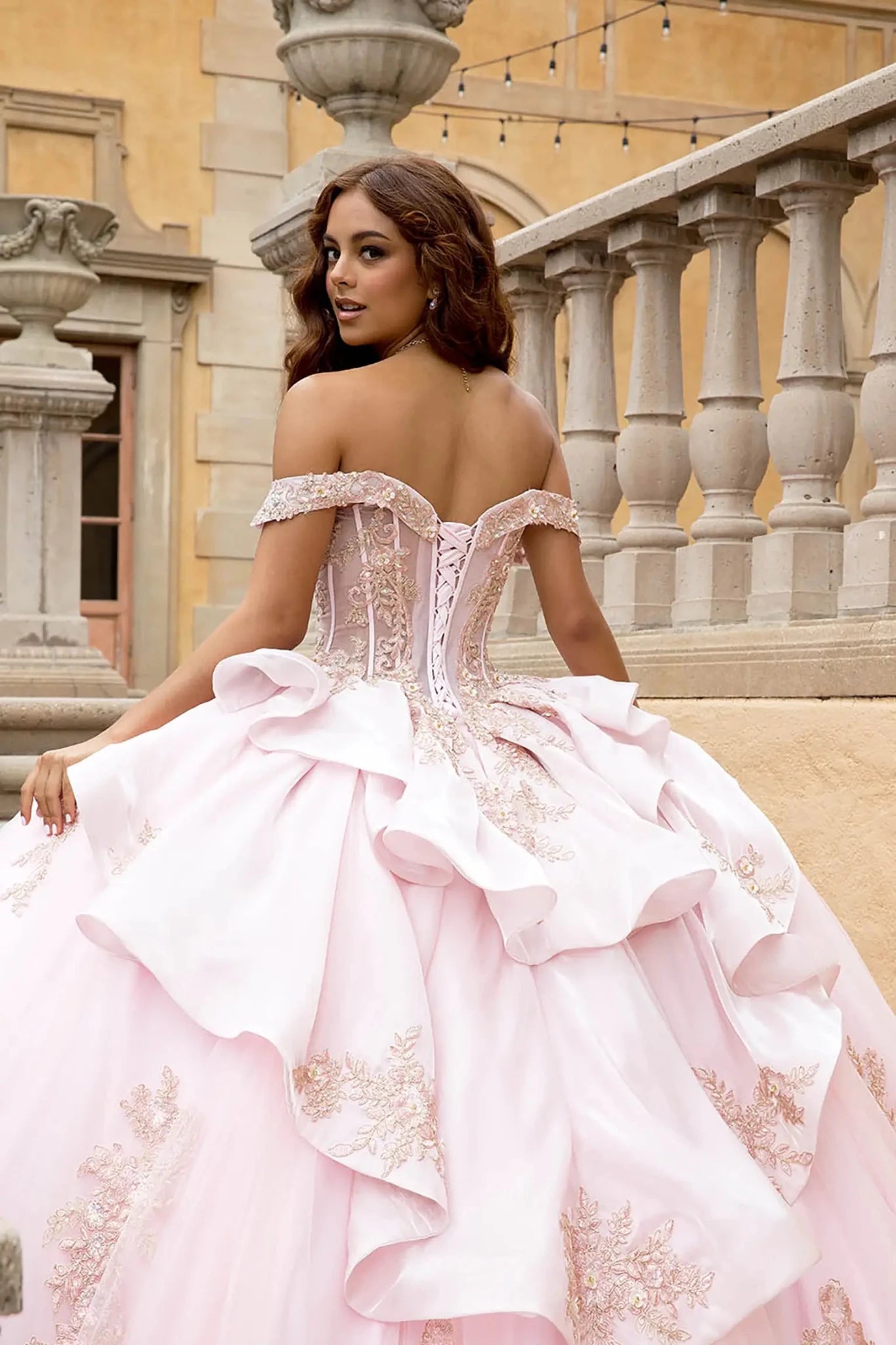 Model shown from behind on a stone balcony, showcasing the sculptural tiered satin ruffles and the adjustable lace-up corset back of the blush quinceañera dress. The view highlights the dramatic volume of the bustle and the continuous floral embroidery on the train.