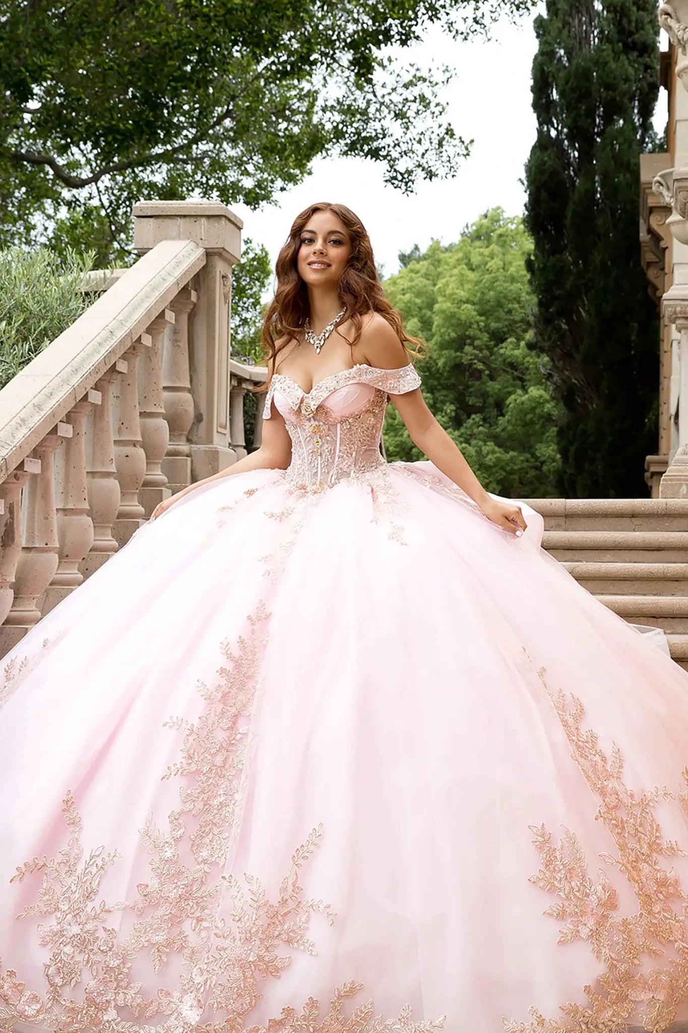 The image captures the sunlight reflecting off the rose gold floral embroidery and sequin accents, emphasizing the regal silhouette of the off-the-shoulder neckline and the massive scale of the glitter tulle ball gown skirt against a luxurious Mediterranean villa background.