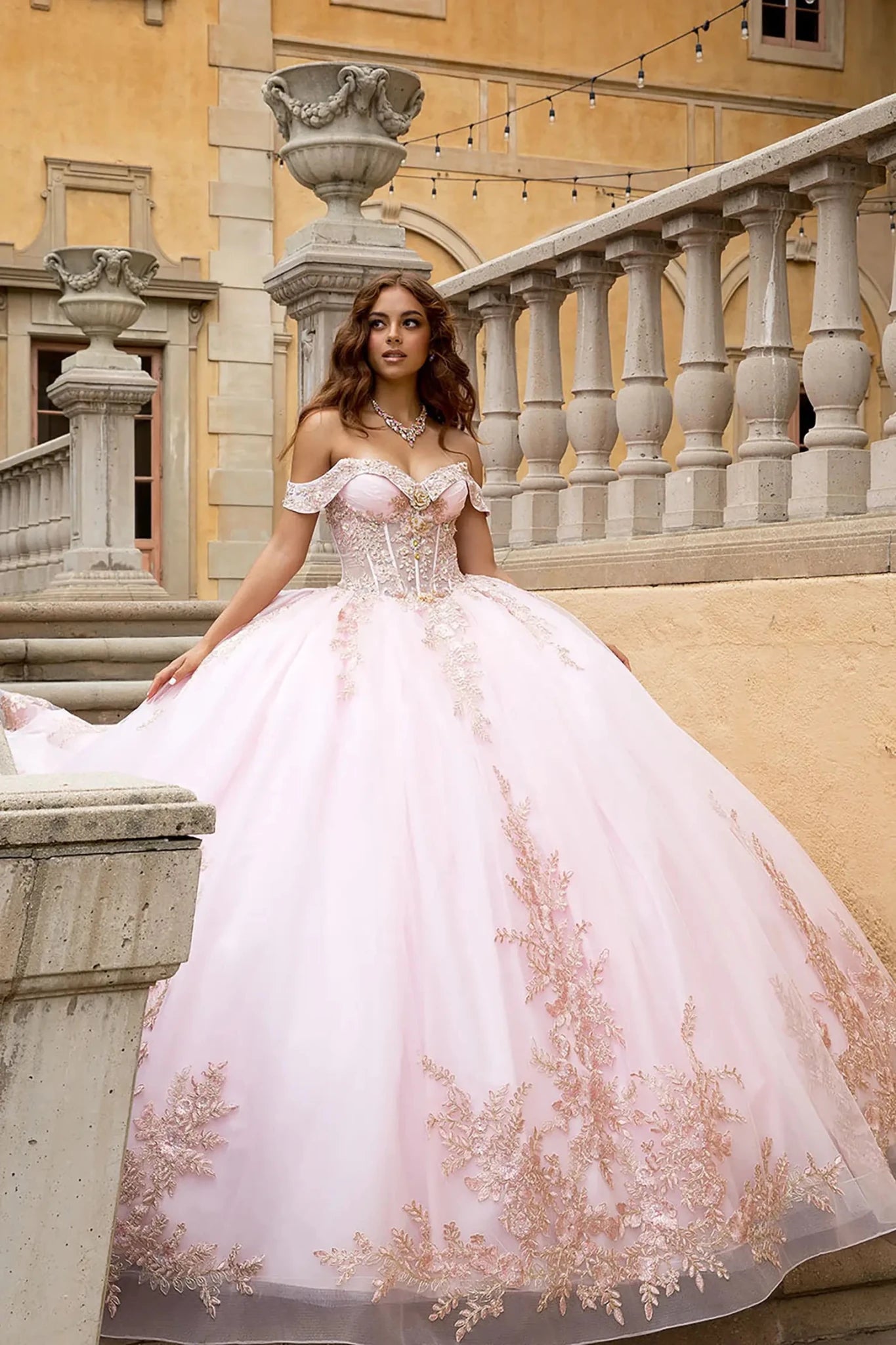 The model is wearing a matching rhinestone statement necklace, set against an elegant Mediterranean villa background.Soft natural sunlight illuminates the fine rose gold embroidery and catches the shimmery, reflective texture of the glitter-infused tulle fabric, creating a luminous glow.