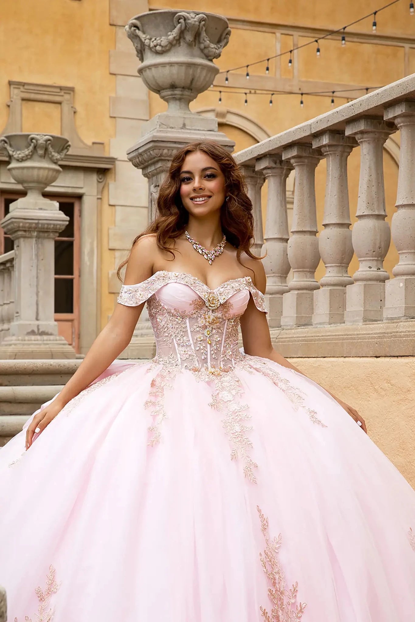 Close up portrait of quinceañera gown on a classical stone staircase. The image highlights the expansive glitter tulle ball gown skirt with rose gold floral embroidery and shimmering lace appliqués cascading toward the hem.