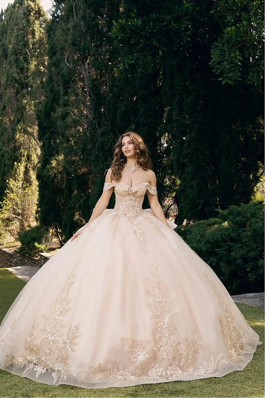 woman standing outdoors on a grassy area with tall, dark green trees in the background. She is wearing an elaborate ball gown in a light blush or champagne color, richly decorated with intricate gold embroidery and embellishments.