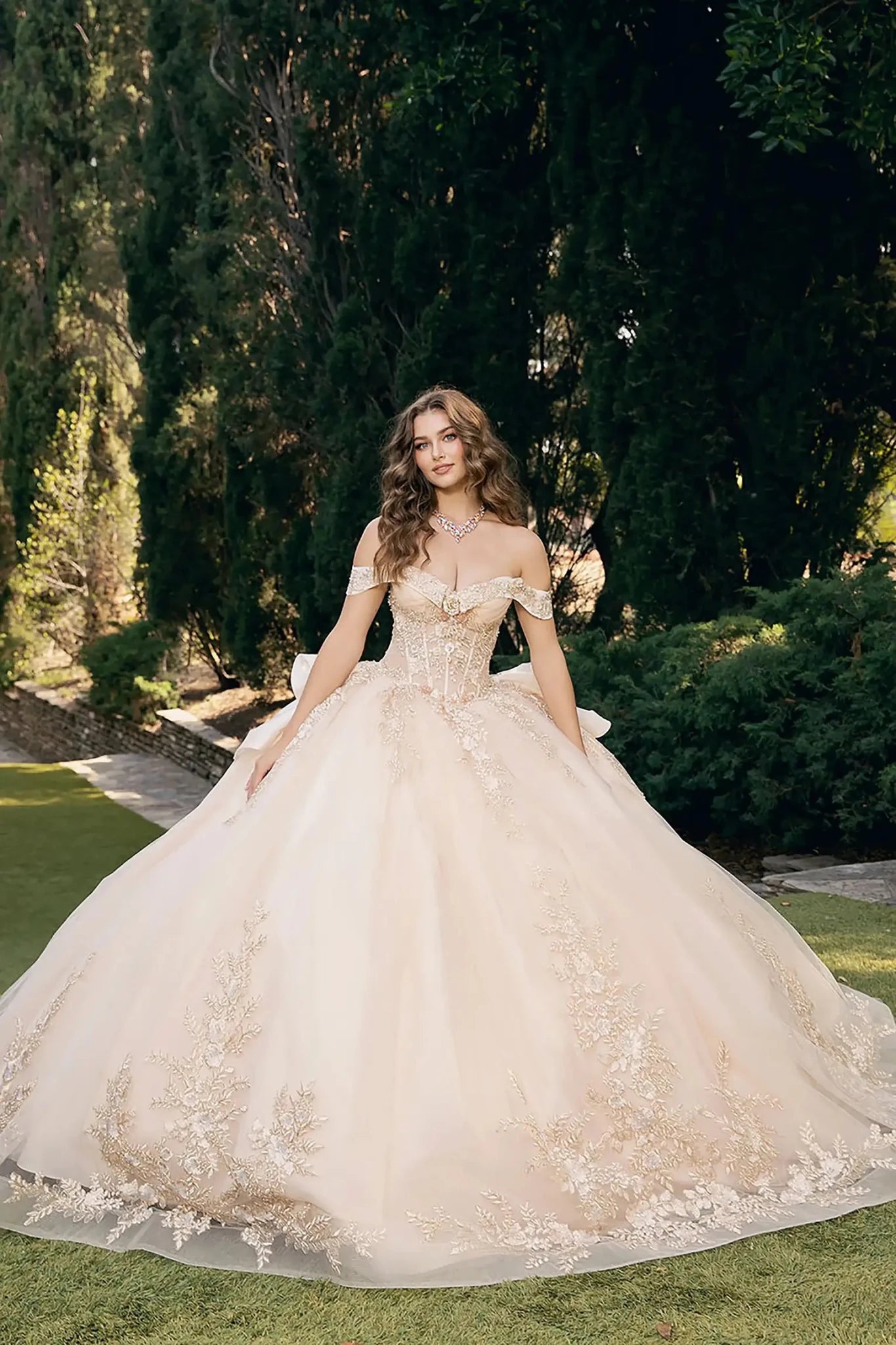 woman standing outdoors in a garden setting with tall, dark green trees and manicured bushes. She is wearing an elaborate off‑the‑shoulder ball gown in a light shade, champagne, richly decorated with intricate floral embroidery and embellishments across the bodice and full skirt.