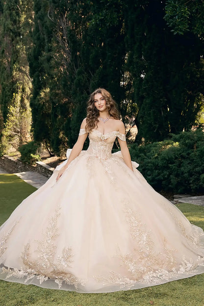 woman standing outdoors in a garden setting with tall, dark green trees and manicured bushes. She is wearing an elaborate off‑the‑shoulder ball gown in a light shade, champagne, richly decorated with intricate floral embroidery and embellishments across the bodice and full skirt.
