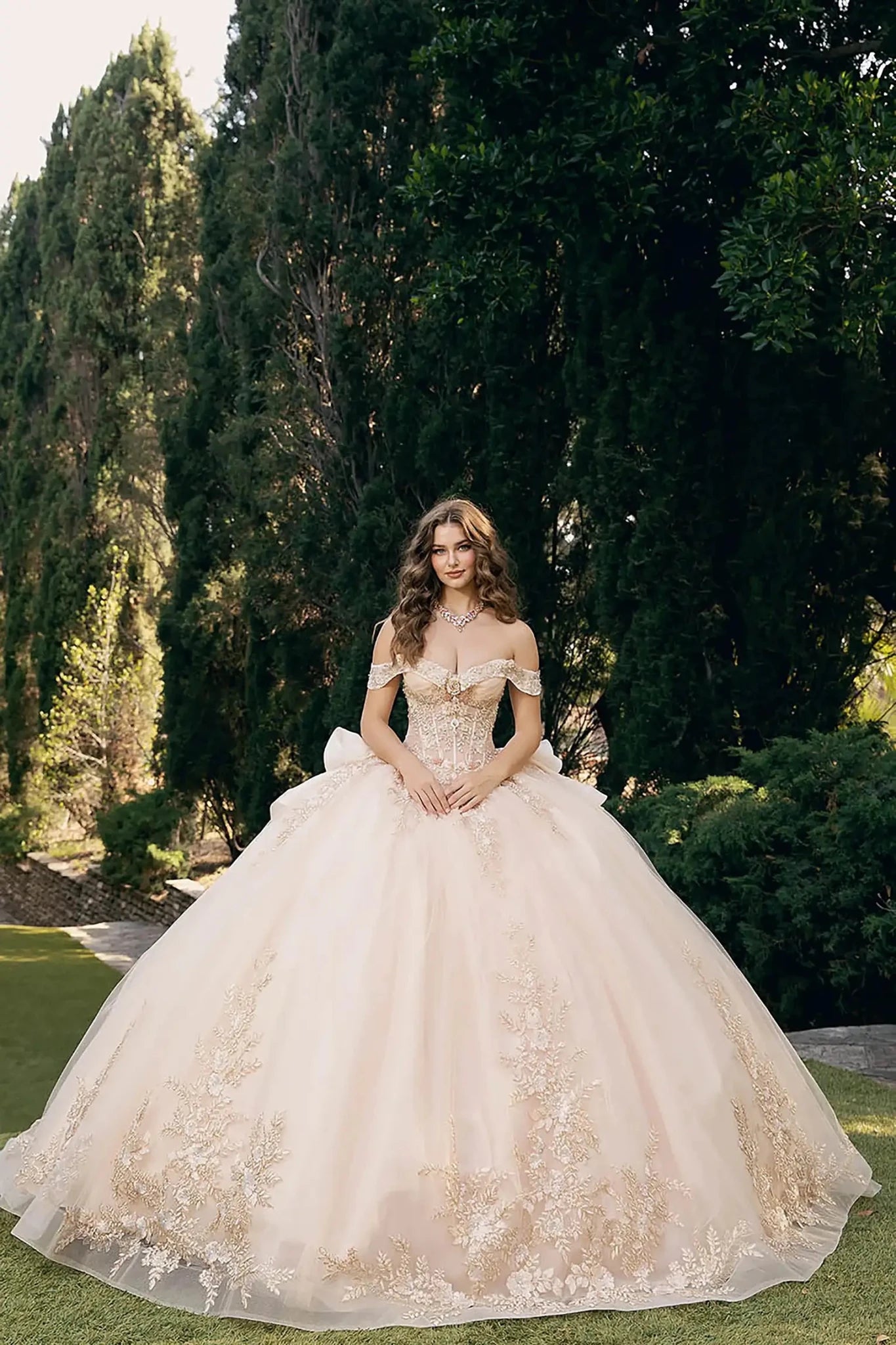 woman standing outdoors in front of tall, lush green trees and manicured bushes. She is wearing an elaborate off‑the‑shoulder ball gown in a light shade, possibly champagne, richly adorned with intricate gold embroidery and lace detailing along the hem and bodice.