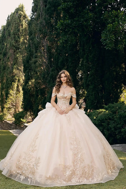 woman standing outdoors in front of tall, lush green trees and manicured bushes. She is wearing an elaborate off‑the‑shoulder ball gown in a light shade, possibly champagne, richly adorned with intricate gold embroidery and lace detailing along the hem and bodice.