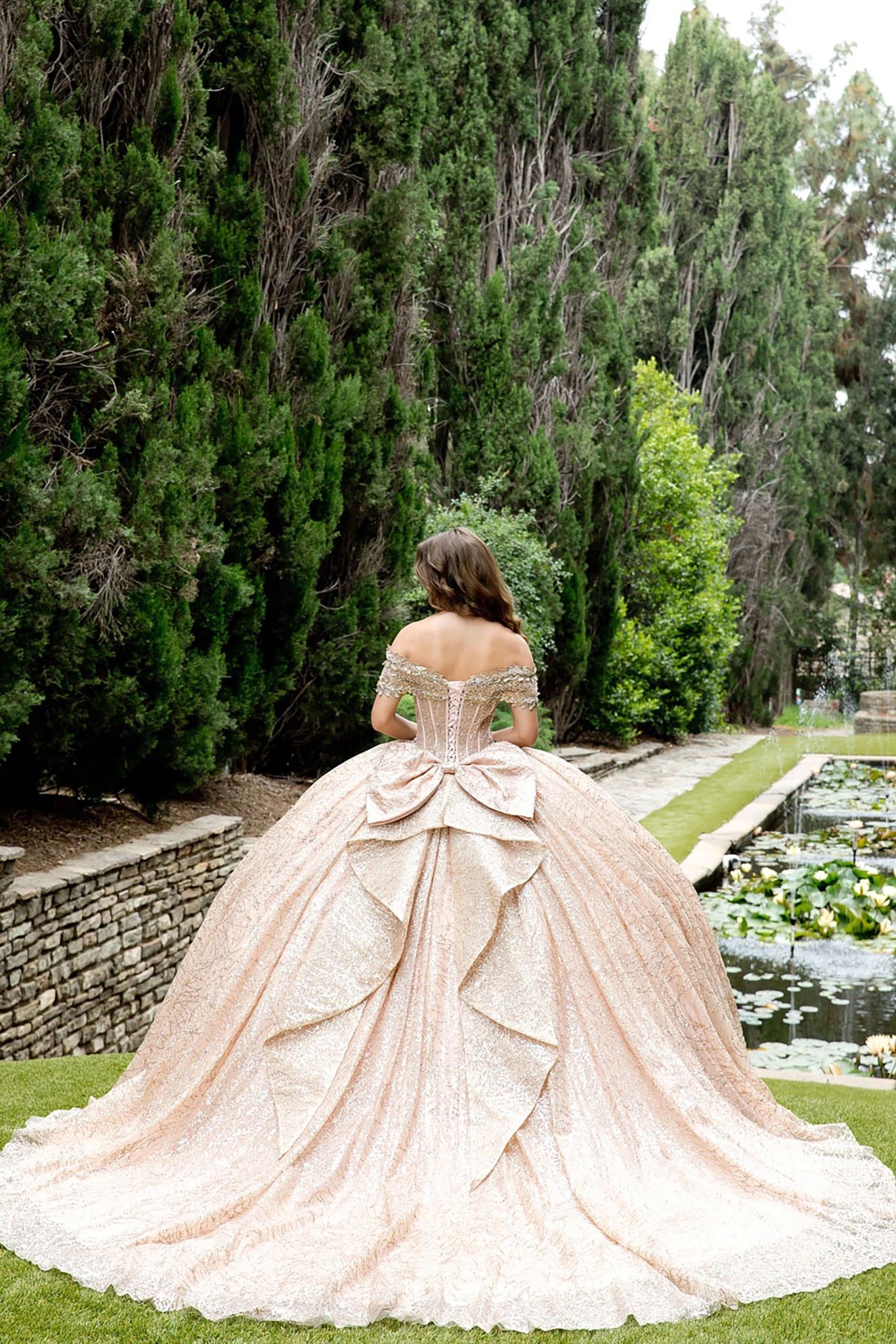 Woman in a rose gold dress standing in a garden with trees and a pond.
