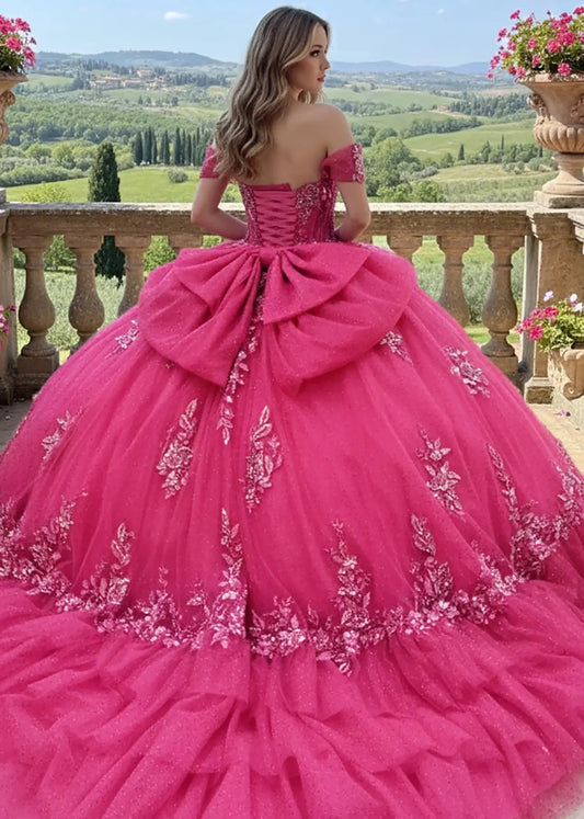 Back view of Fuchsia off-shoulder ball gown with lace-up corset back, oversized bow detail, and glitter floral appliqué skirt, perfect for quinceañera