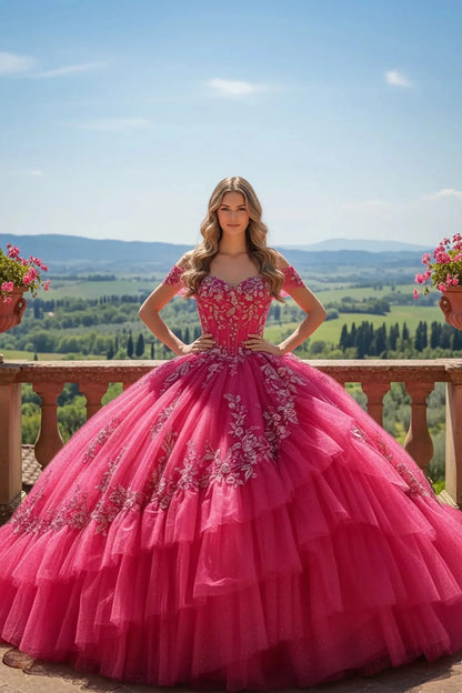 Woman in a vibrant, fuchsia-pink ball gown with intricate silver-embroidered details stands on a stone balcony, overlooking a picturesque landscape of rolling green hills and distant mountains under a clear blue sky. She has long, wavy blonde hair and a serene expression, with her hands resting on her hips, showcasing the voluminous, tiered skirt of her dress. Two terracotta pots with pink flowers are visible on either side of her on the balcony railing.