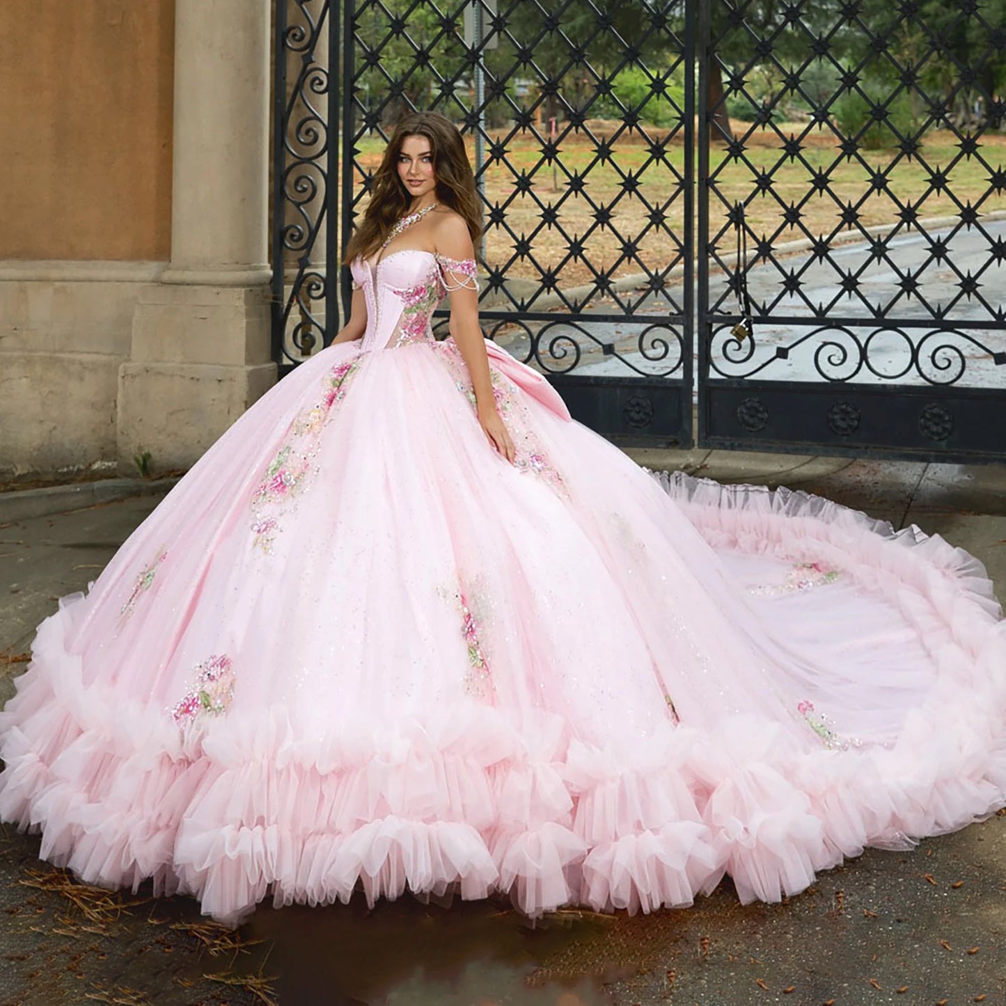 Pink ball gown with floral details worn by a model in front of an ornate gate.