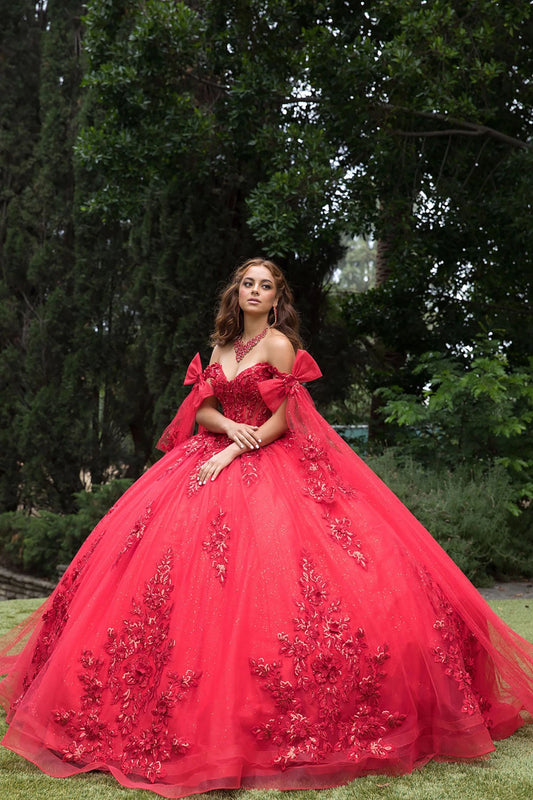 Woman in a red ball gown with floral details standing outdoors among trees.