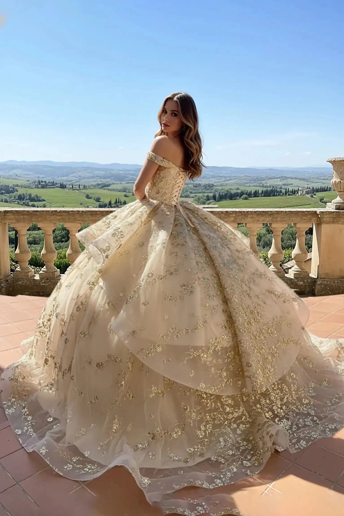champagne ballgown with an off-the-shoulder neckline and a ruffled peplum waist, featuring intricate gold floral lace embroidery posing on a vineyard balcony.
