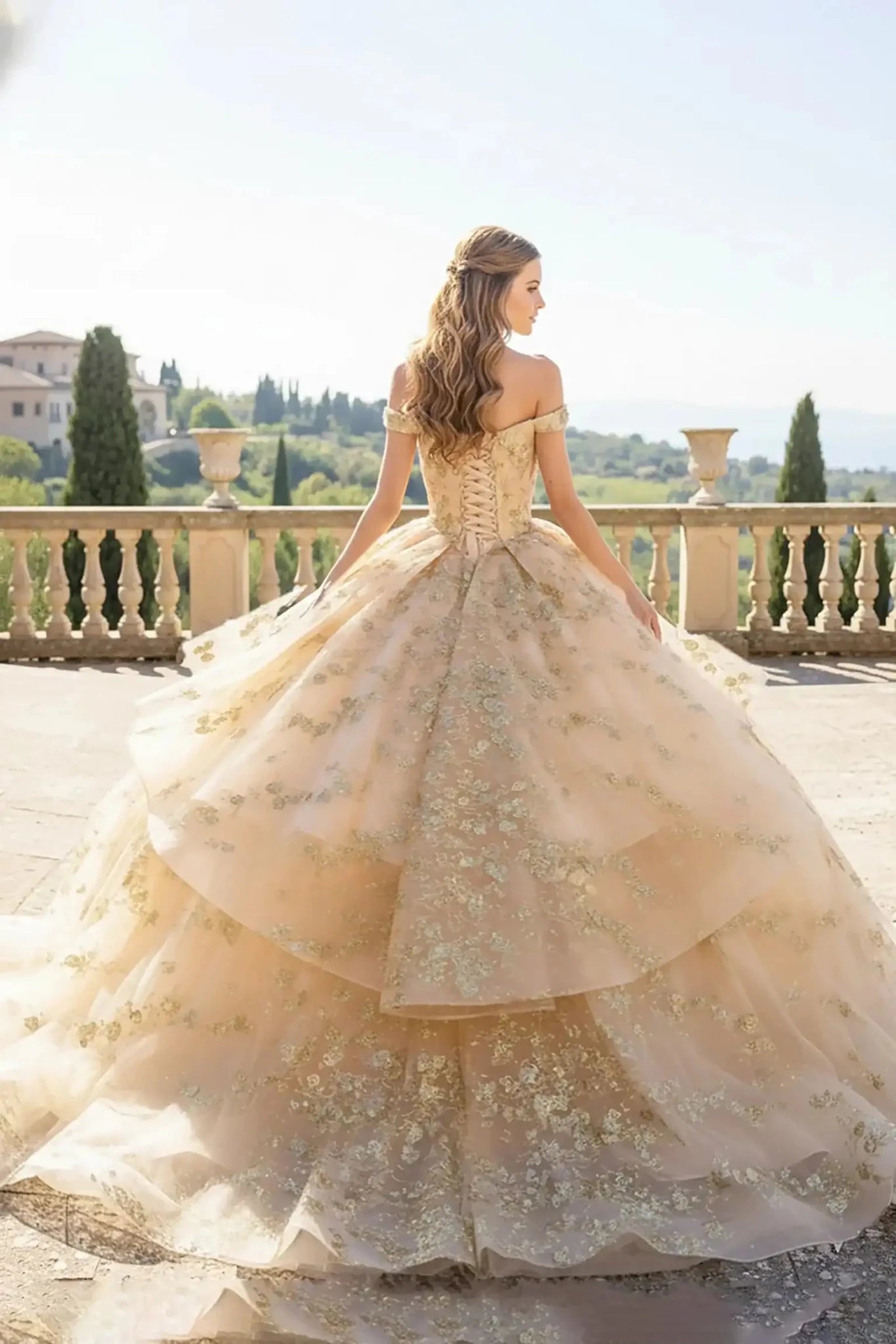 back view champagne ballgown with an off-the-shoulder neckline and a ruffled peplum waist, featuring intricate gold floral lace embroidery posing on a vineyard balcony.