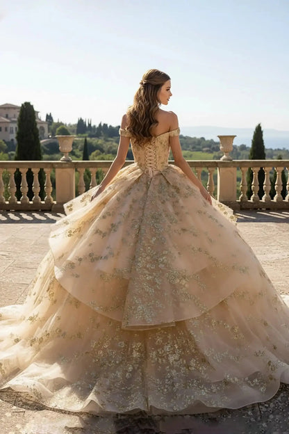 Back of full-length champagne ballgown with an off-the-shoulder neckline and a ruffled peplum waist, featuring intricate gold floral lace embroidery posing on a vineyard balcony.