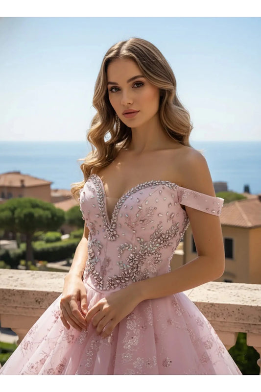 Half view of a pink ballgown with an off-the-shoulder neckline and a tiered peplum waist, decorated with shimmering floral embroidery and crystal accents against a coastal sea horizon.