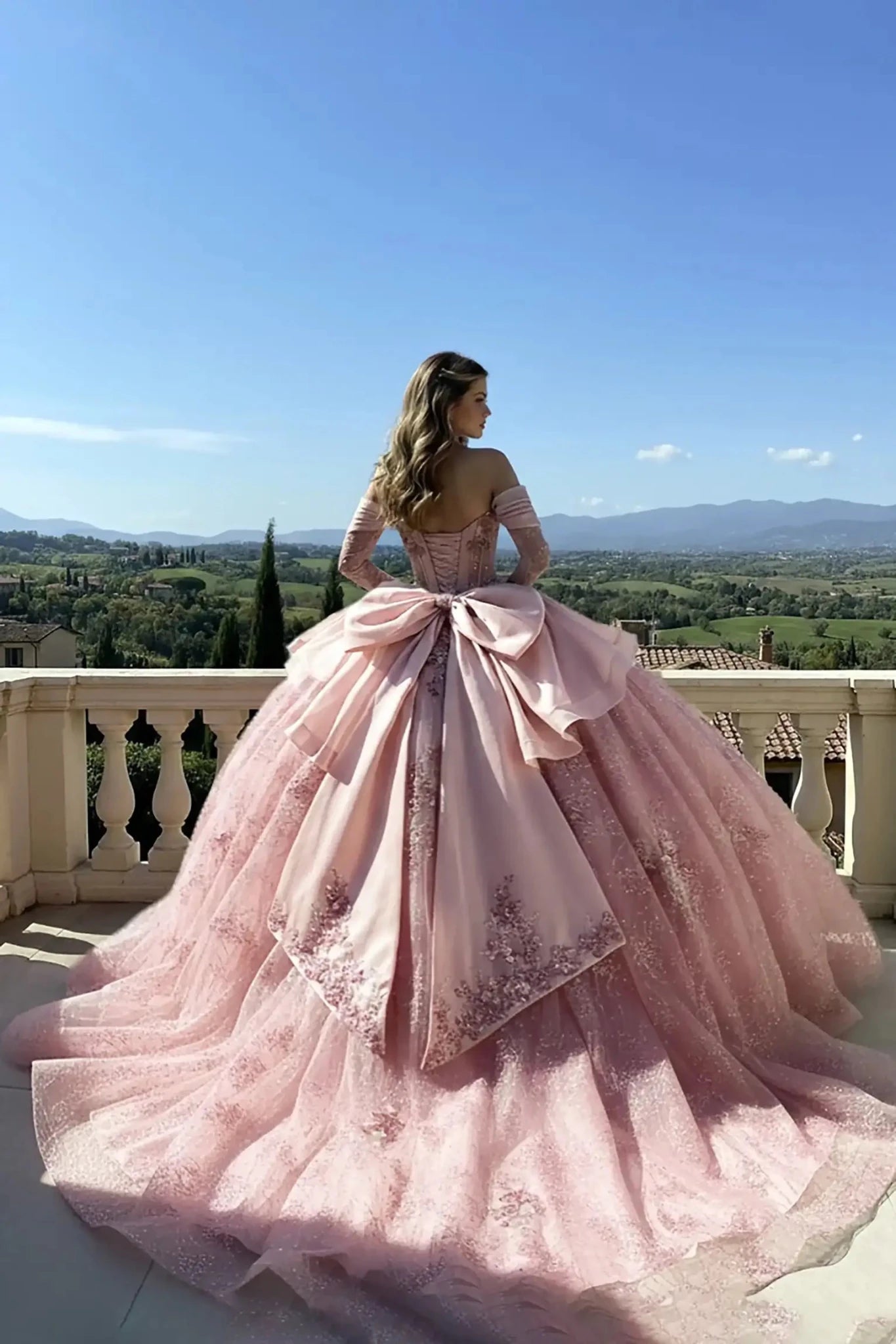 Rear view of a pink off-the-shoulder ball gown featuring a corset-style back, a large decorative bow, and an expansive floral embroidered train, set on a stone terrace with tall cypress trees in the background.