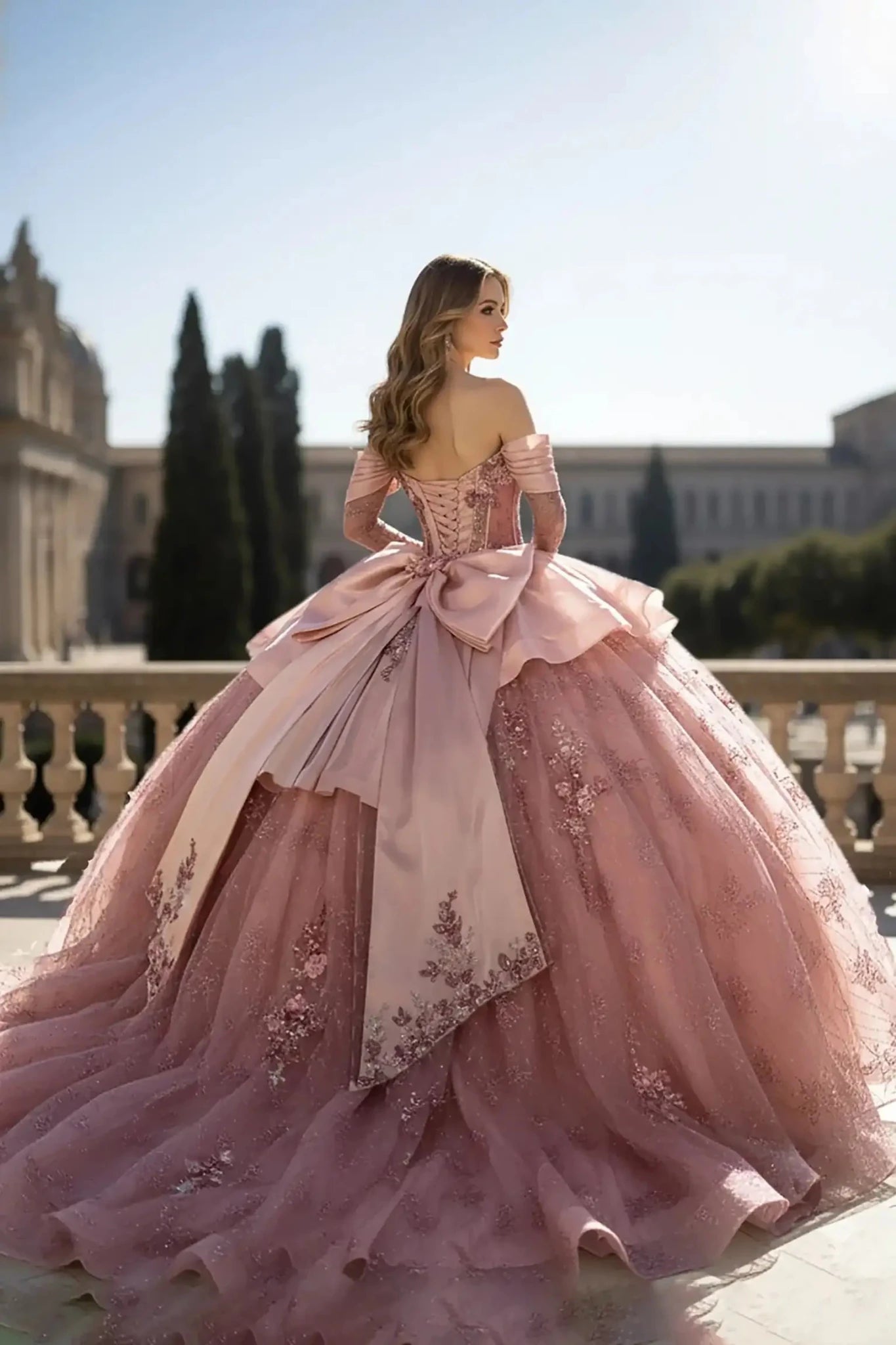 Back view of a blush pink off-the-shoulder ball gown featuring a corset-style back, a large decorative bow, and an expansive floral embroidered train, set on a stone terrace with tall cypress trees in the background.