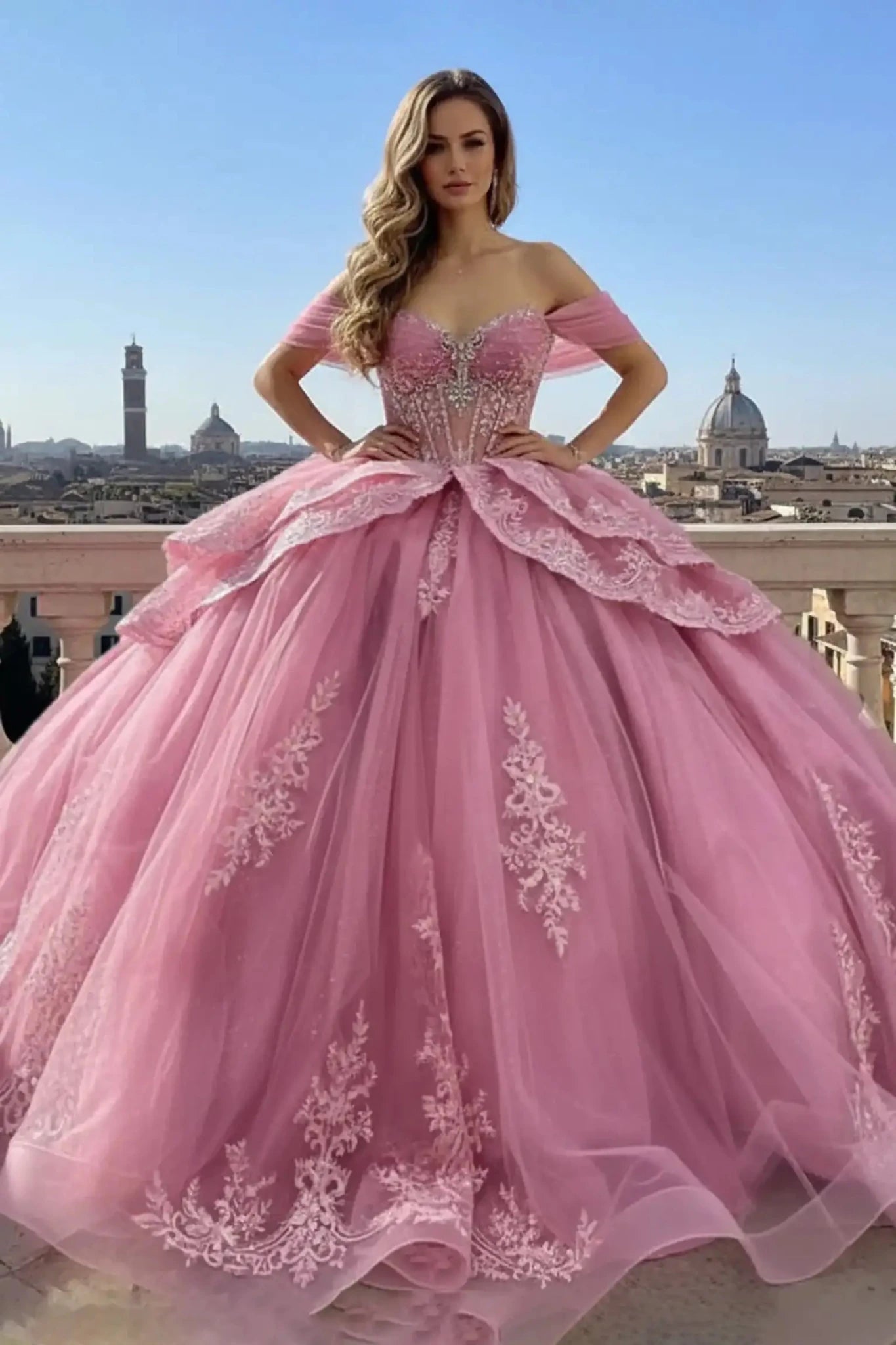 Full length of pink ballgown with an off-the-shoulder sweetheart neckline, featuring silver lace appliqués on the corset and skirt, shown on a model with a city skyline and dome architecture background.