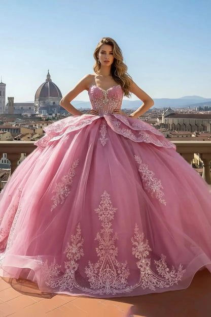 Front view of a rose pink ballgown featuring a lace-up back, large bow, and tiered lace-trimmed bustle, shown against a panoramic European city view with historical cathedrals.