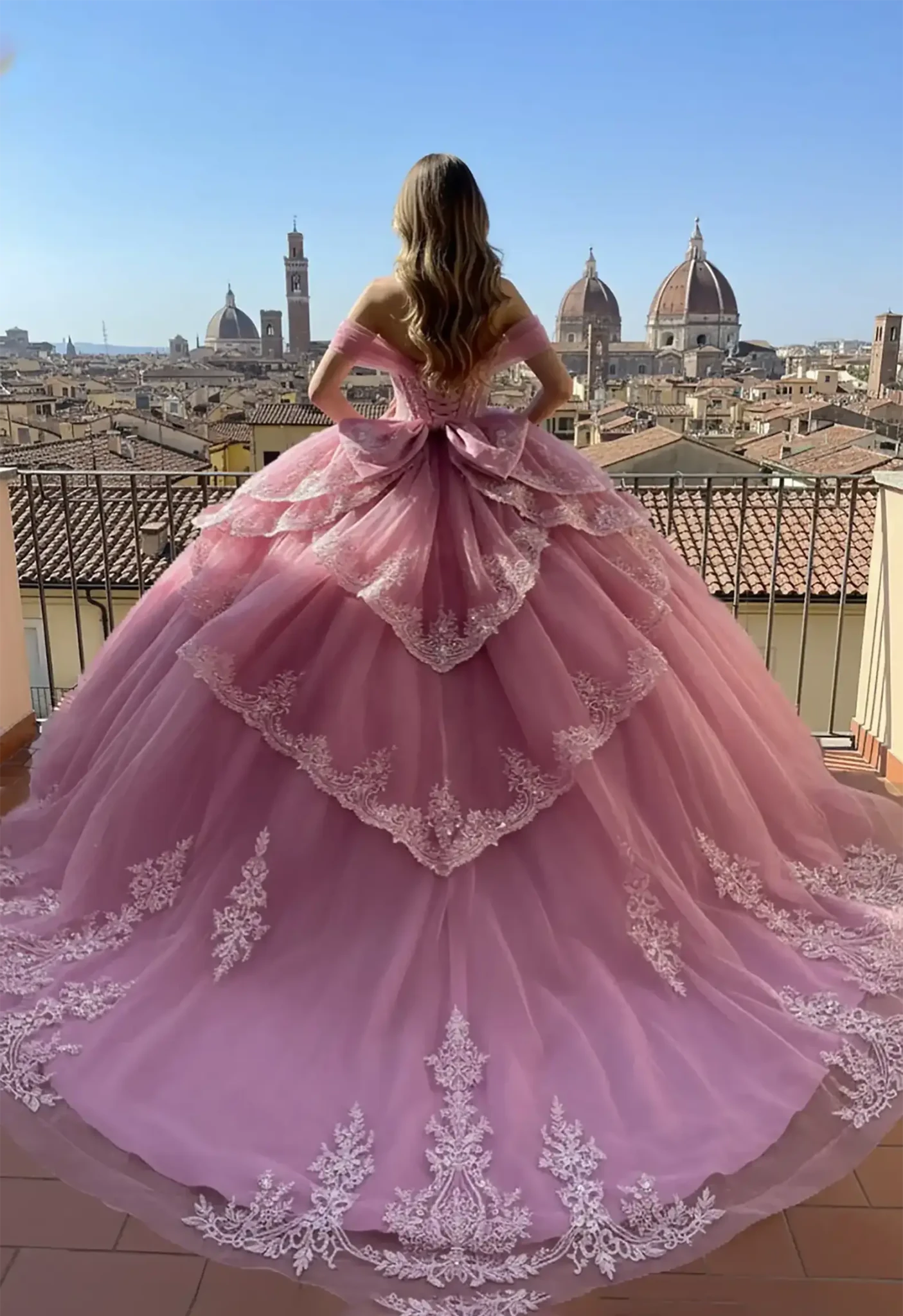 Back view of a pink ballgown featuring a lace-up back, large bow, and tiered lace-trimmed bustle, shown against a panoramic European city view with historical cathedrals.