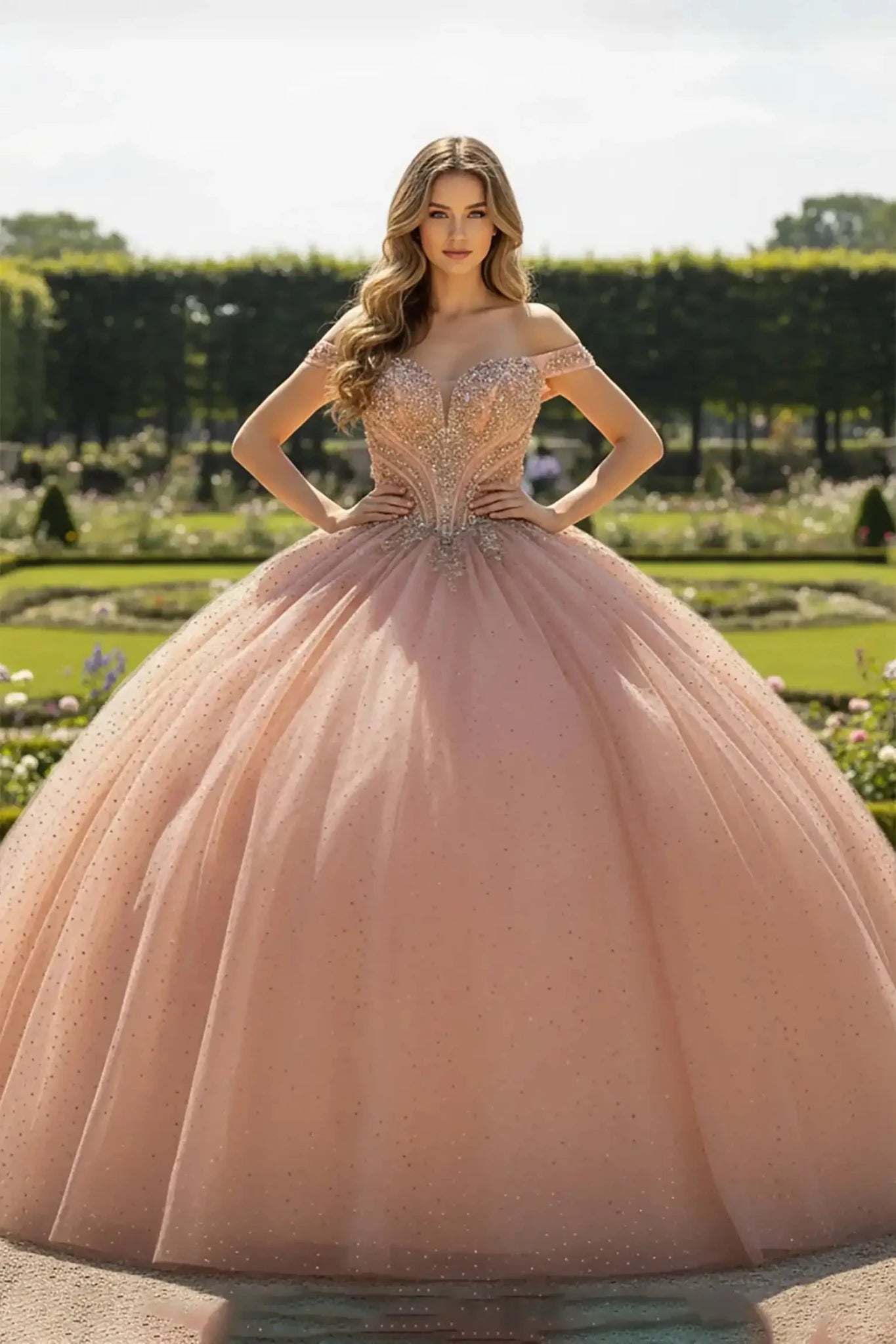 Rose Gold ballgown with a glittering tulle skirt and off-the-shoulder sweetheart bodice, decorated with shimmering crystal beadwork, shown in a manicured garden setting.