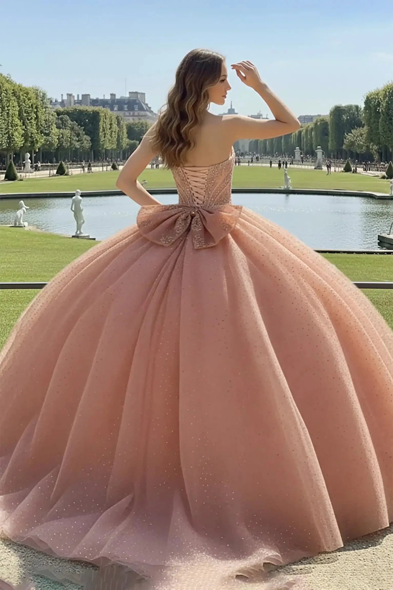 Back view of a Rose Gold glitter ballgown with a lace-up corset and a large decorative bow, showing a voluminous sparkling tulle skirt in a park setting with a pond.