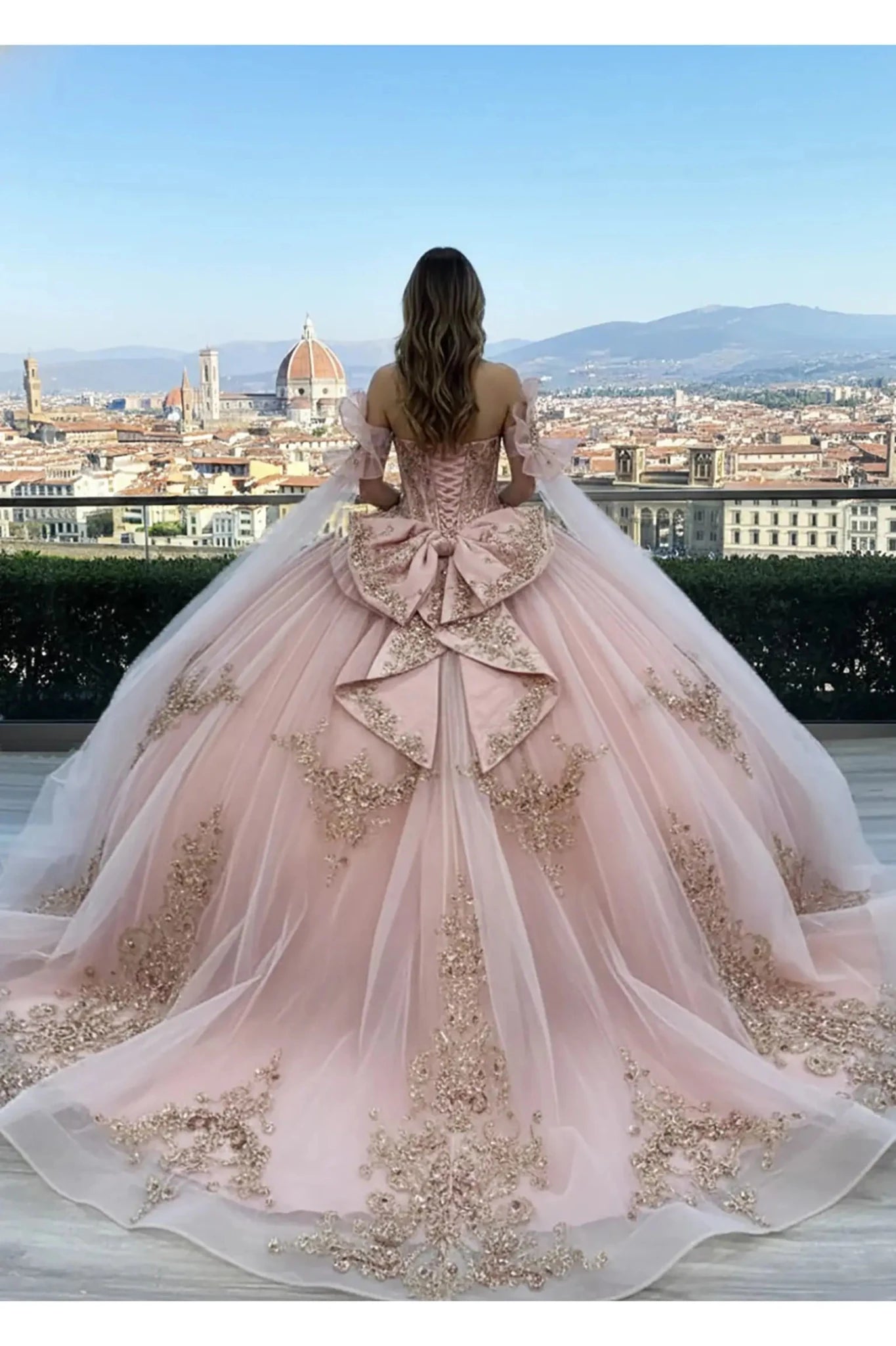Back view of a Rose Gold off-the-shoulder ball gown featuring a corset-style lace-up back, a voluminous decorative bow at the waist, and an expansive floor-length train with delicate floral embroidery, set on a stone terrace with tall cypress trees in the background.