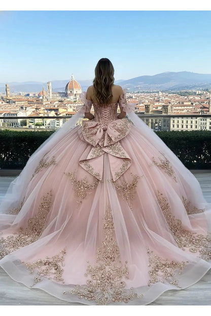 Back view of a Rose Gold off-the-shoulder ball gown featuring a corset-style lace-up back, a voluminous decorative bow at the waist, and an expansive floor-length train with delicate floral embroidery, set on a stone terrace with tall cypress trees in the background.