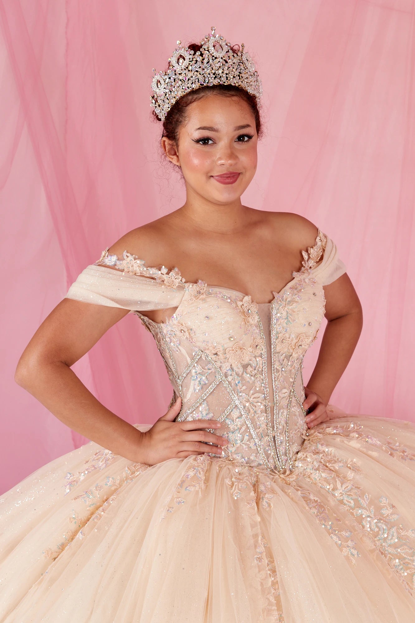 Woman in a champagne off-shoulder gown with floral details and a tiara against a pink background
