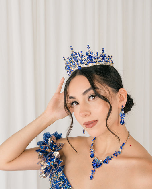 Woman wearing a royal blue tiara and matching jewelry set against a white curtain background