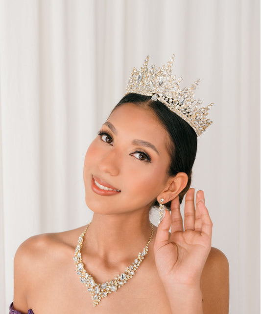 Woman wearing a gold bold tiara and necklace, holding her hand to her ear, against a plain background