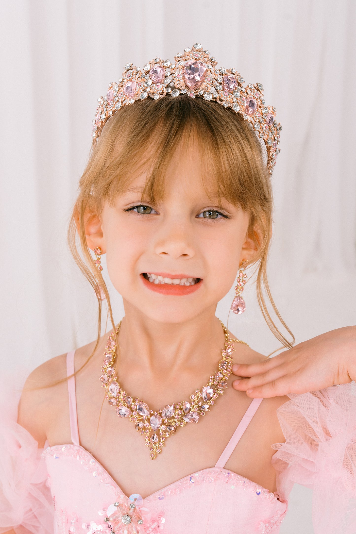 Young girl wearing a pink dress, tiara, and necklace against a white background