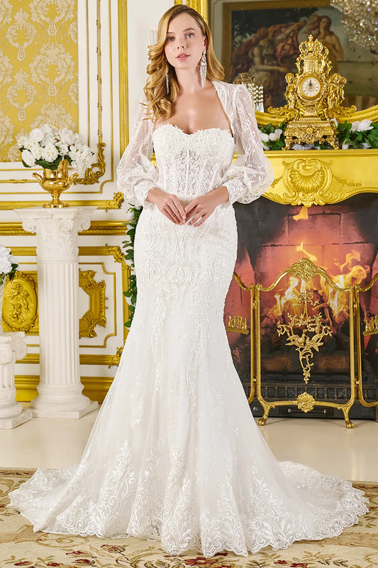 A girl wearing a trumpet-shaped wedding dress fully adorned with luxurious lace. She is standing in a sleeveless wedding gown layered with a long-sleeve lace bolero. The skirt flares out beautifully toward the hem and is finished with wide lace detailing