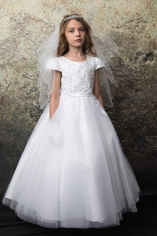 Young girl in a white lace dress with a veil against a textured stone wall.