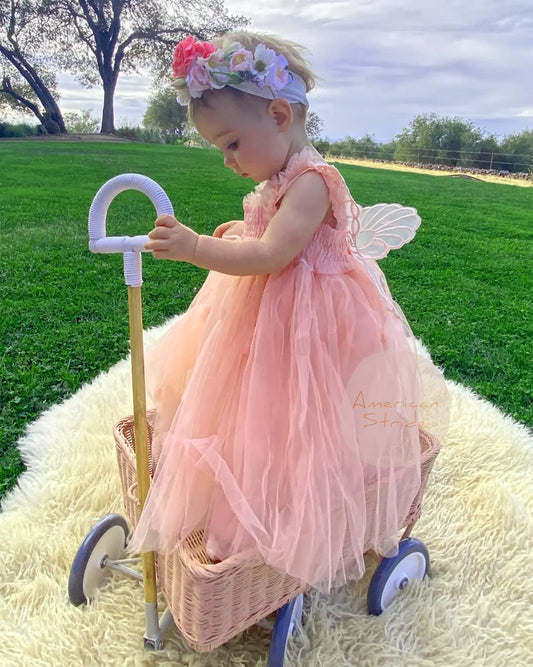 Child in a pink dress with fairy wings and a flower crown, standing next to a toy basket and walking stick outdoors.