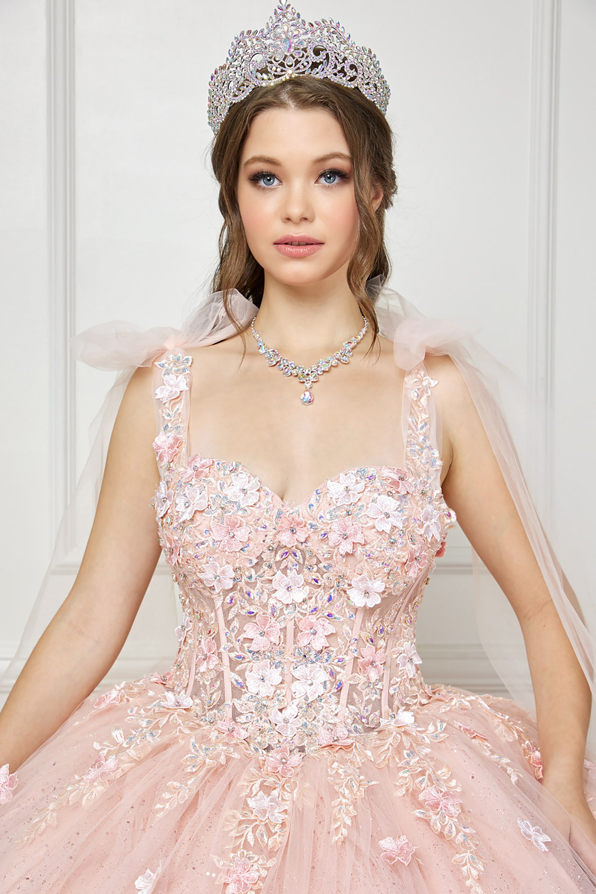 Quinceanera | Blush-Peach Flower Embroidery, Gem Detailing, and glitter lining Quince Ballgown with Tulle Shoulder Drape - zoomed in view of woman wearing dress and crown looking into the camera