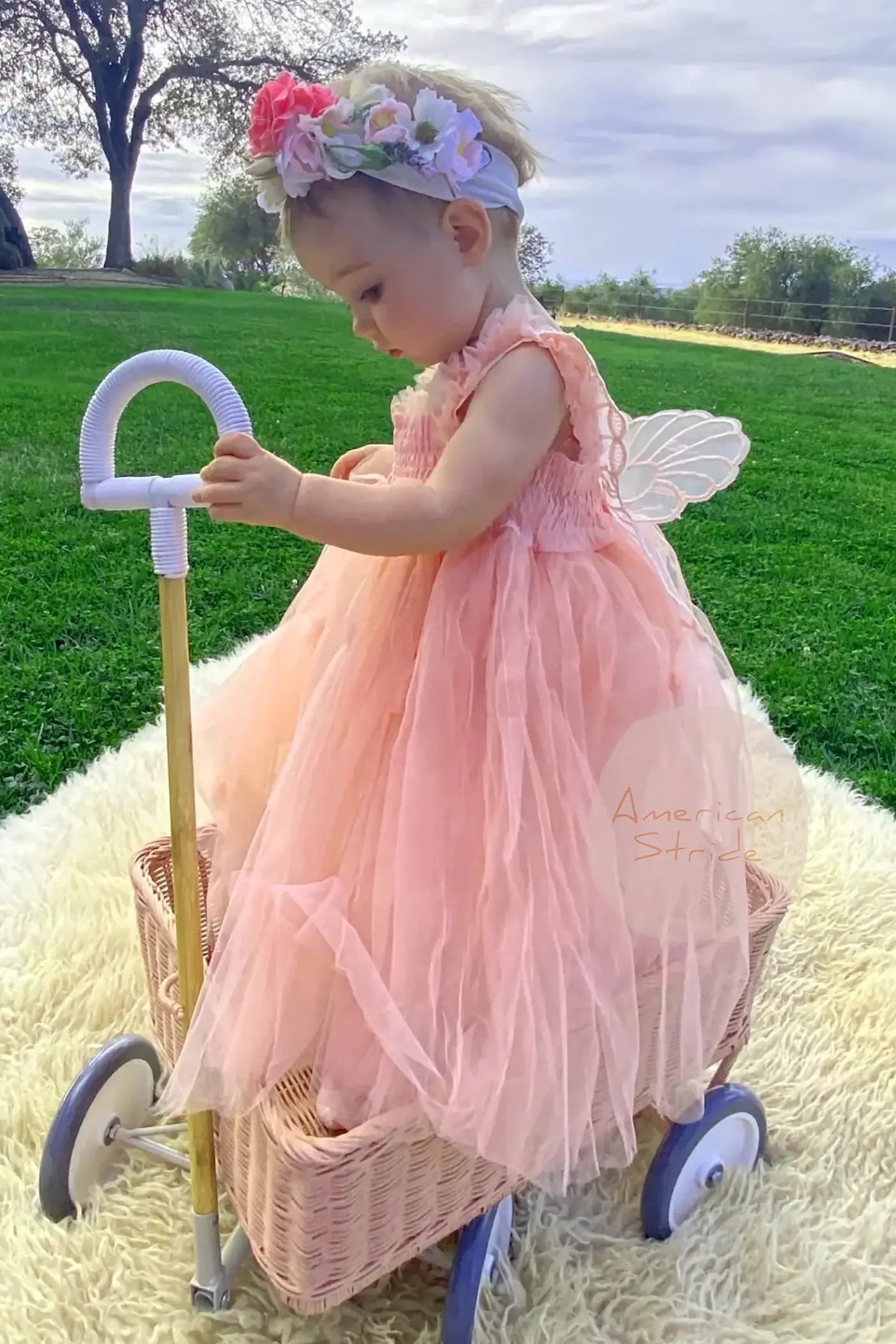 Child in a pink tulle dress with fairy butterfly wings standing next to a toy stroller outdoors.