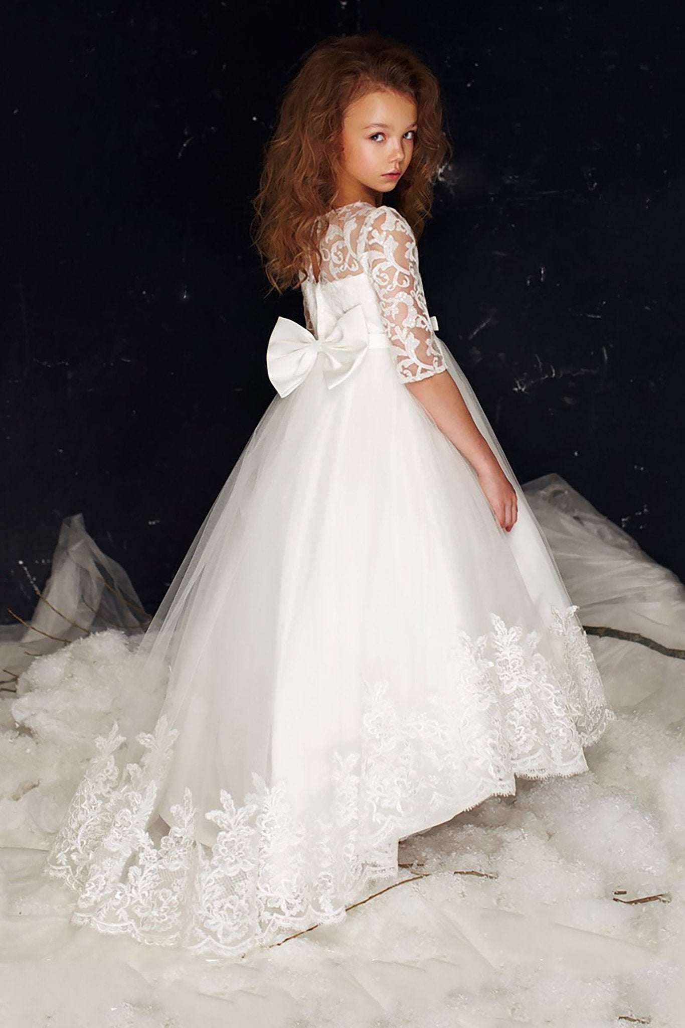 Three-quarter back view of a white communion dress with three-quarter floral lace sleeves, a satin waist bow, and a lace-trimmed high-low tulle skirt.