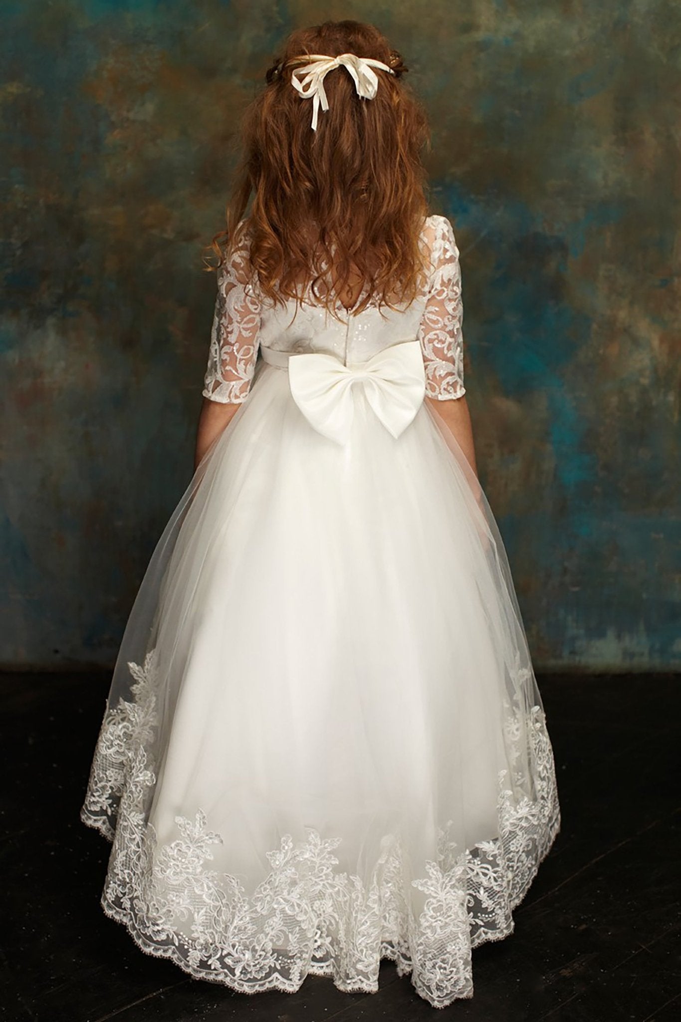 Back view of a white communion dress with three-quarter floral lace sleeves, a large satin bow, and a lace-trimmed tulle skirt.