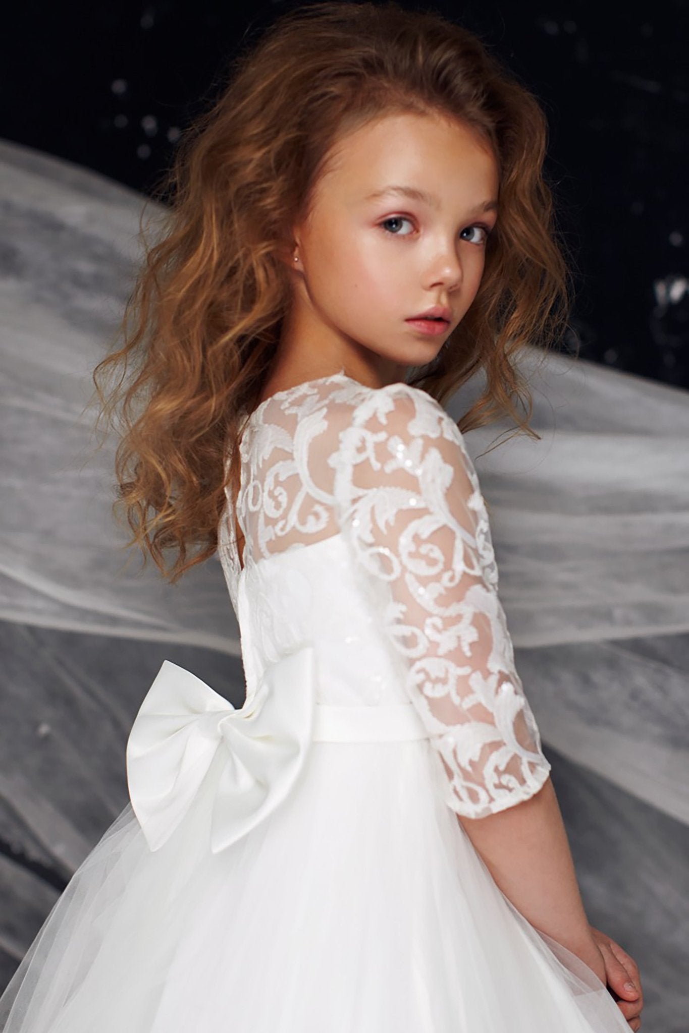 Close-up back view of a white communion dress showing a floral lace sleeve and a large satin waist bow.