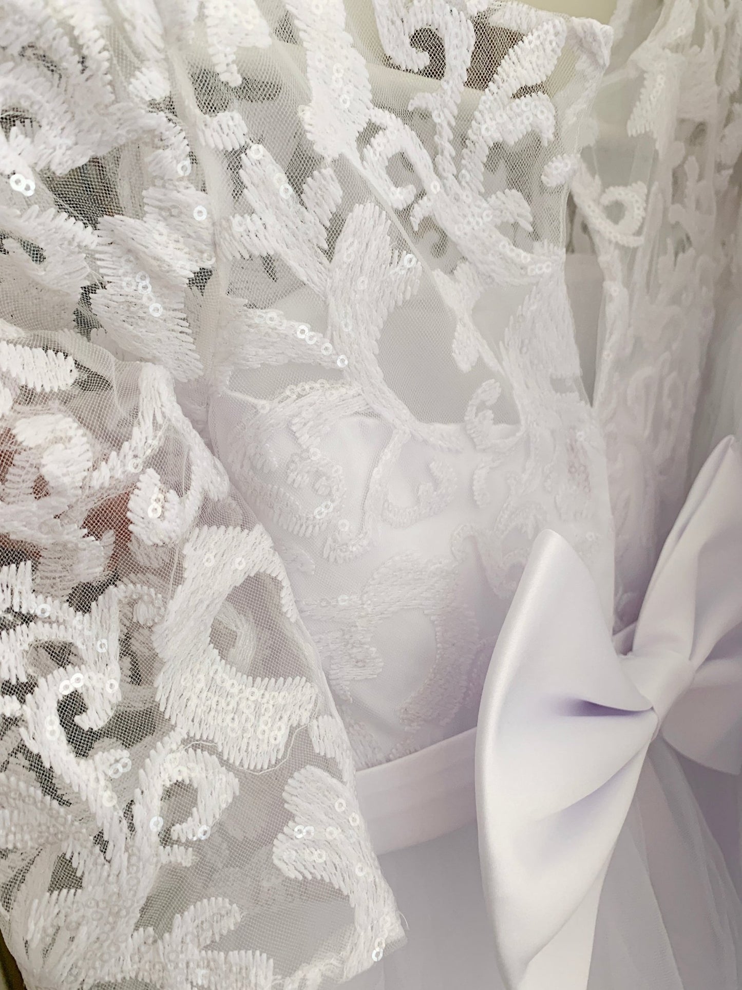 Close-up of white floral lace with sequin details beside a satin bow on a communion dress.