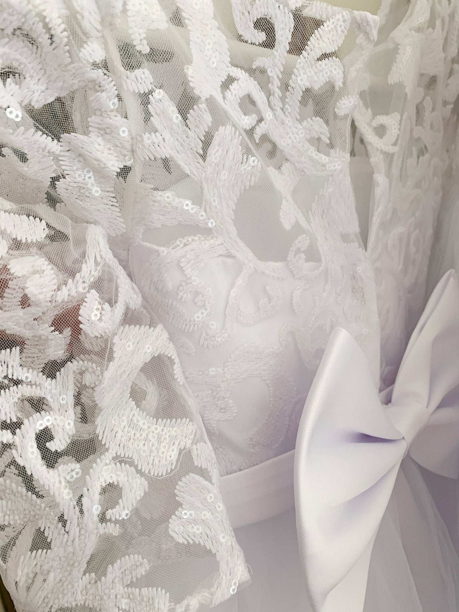 Close-up of white floral lace with sequin details beside a satin bow on a communion dress.